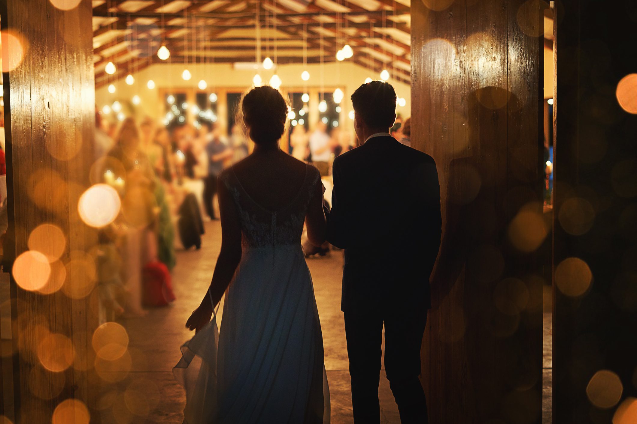 Couple walking hand in hand through warmly lit wedding reception.