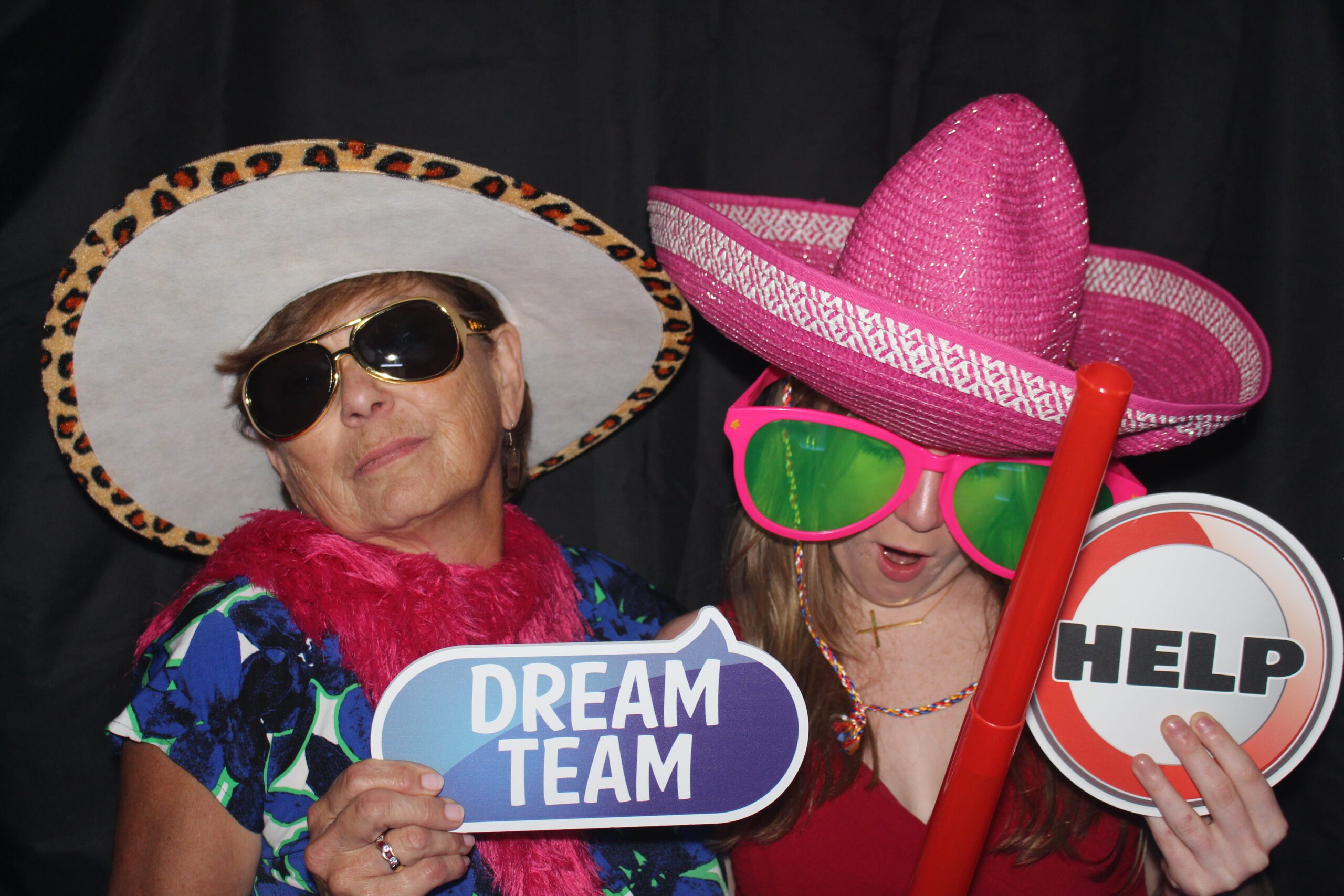 Two women in colorful hats and sunglasses posing with playful signs.